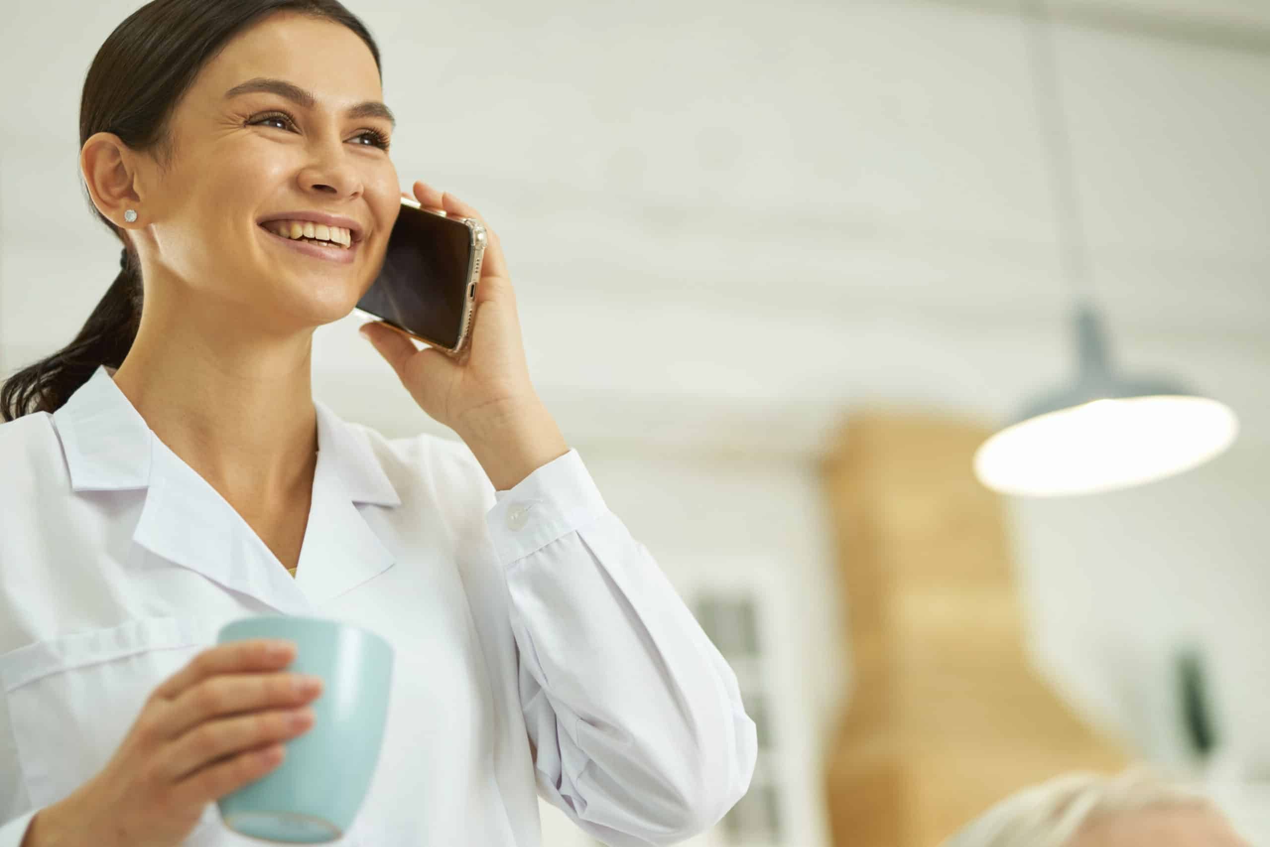 Smiling female doctor using smartphone and drinking hot drink